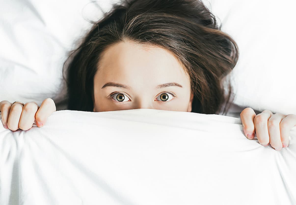 Une jeune femme se cache le visage derrière un drap de lit.